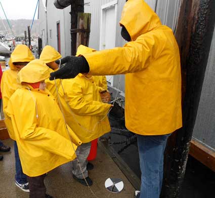 An instructor directs youth students to a dock. They are all wearing bright yellow rain jackets.