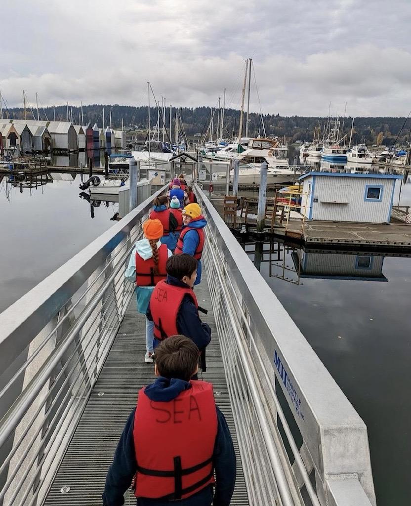 Group of children in life jackets walking on a marina dock, surrounded by boats and cloudy skies.